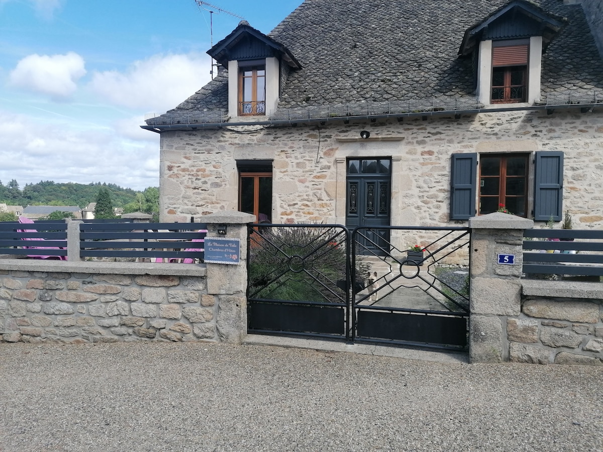 A stone-faced exterior of a traditional house is presented, featuring dark window shutters and a central door. A black wrought iron gate opens into a gravel path. Flowering plants can be seen alongside the stone wall, enhancing the entrance.