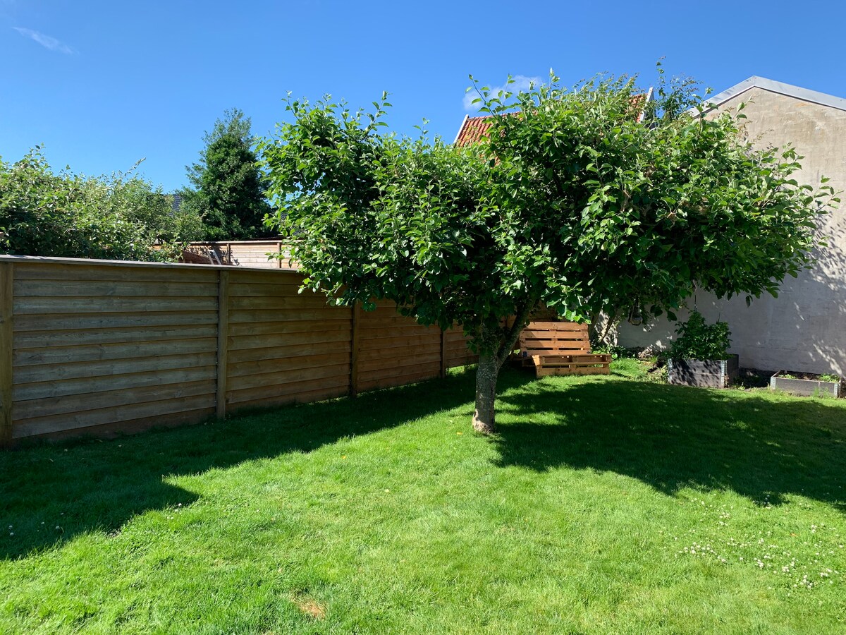 The spacious garden features a well-maintained green lawn, complemented by a sturdy wooden fence. A leafy tree stands prominently, providing shade. In the background, a wooden seating area can be seen, offering a relaxing spot amidst the natural surroundings.