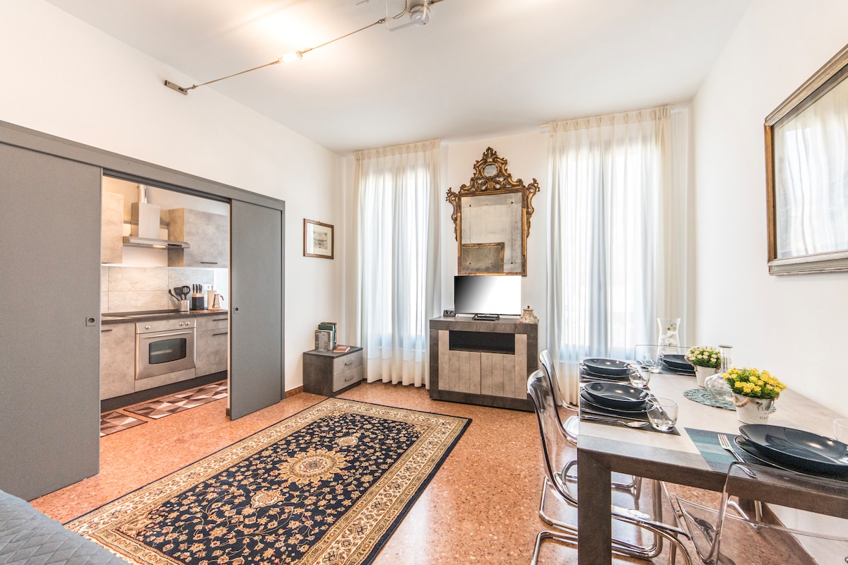 A well-lit living space features large windows draped with light curtains, allowing natural light to fill the room. A dining table with place settings is visible alongside a decorative mirror and a small decorative plant. The kitchen area is partially shown, featuring modern appliances.