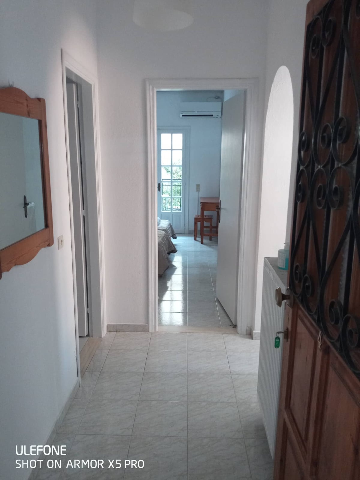 A bright hallway is visible, featuring tiled flooring and white walls. Doors lead to the bedrooms on one side, while a mirror reflects light and creates a sense of openness. Sunlight enters from a window at the end, illuminating the space.