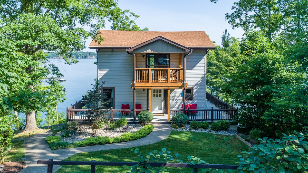 The home features a two-story structure surrounded by lush greenery and well-maintained landscaping. A path leads to the front entrance, flanked by flower beds and shrubs. The wrap-around deck includes seating areas, and large windows provide views of the lake.