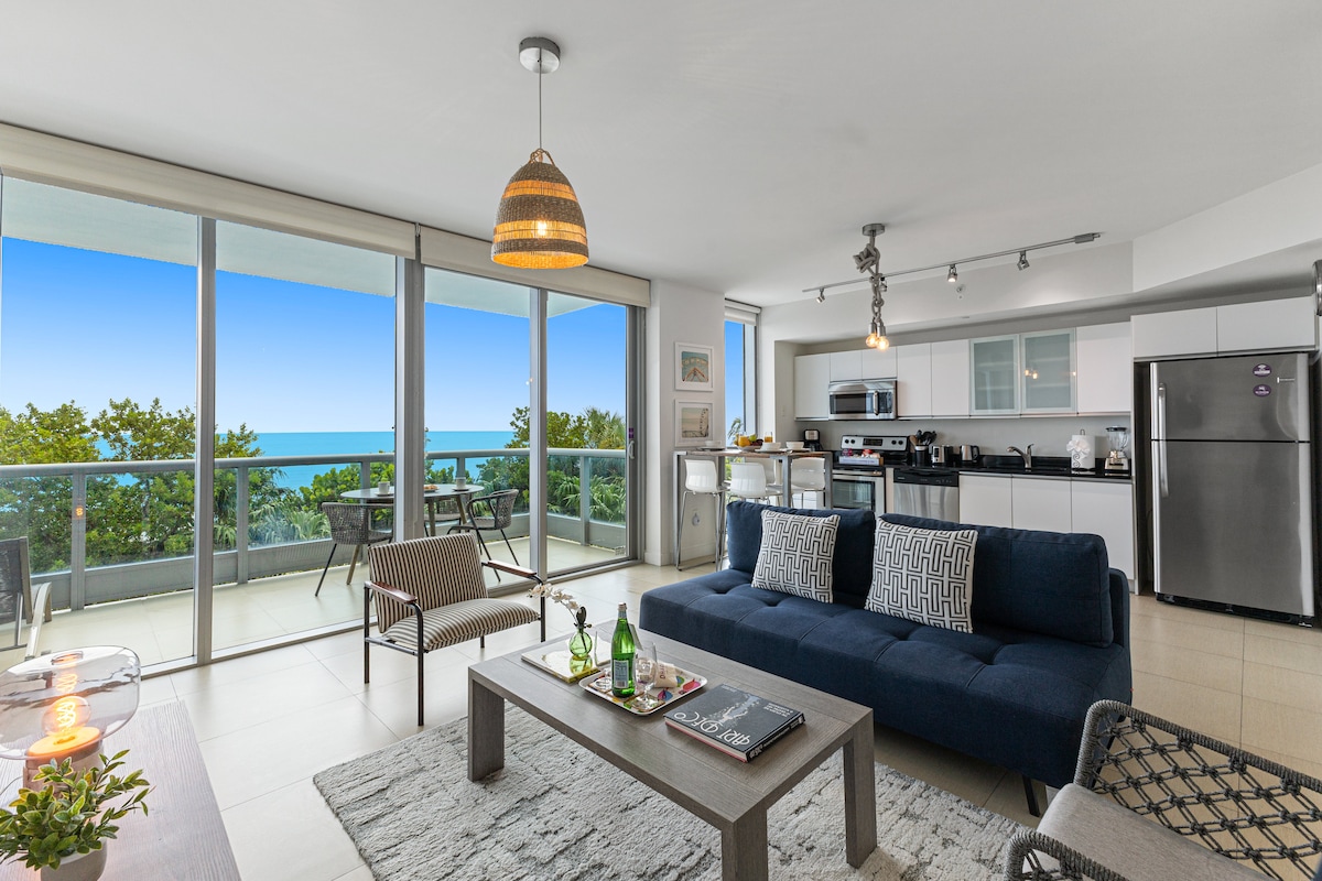 A modern living area is featured with a spacious sofa and a coffee table. Large glass doors provide an unobstructed ocean view and access to a private balcony. A kitchen with stainless steel appliances is visible, along with cozy seating arrangements, creating a welcoming space.