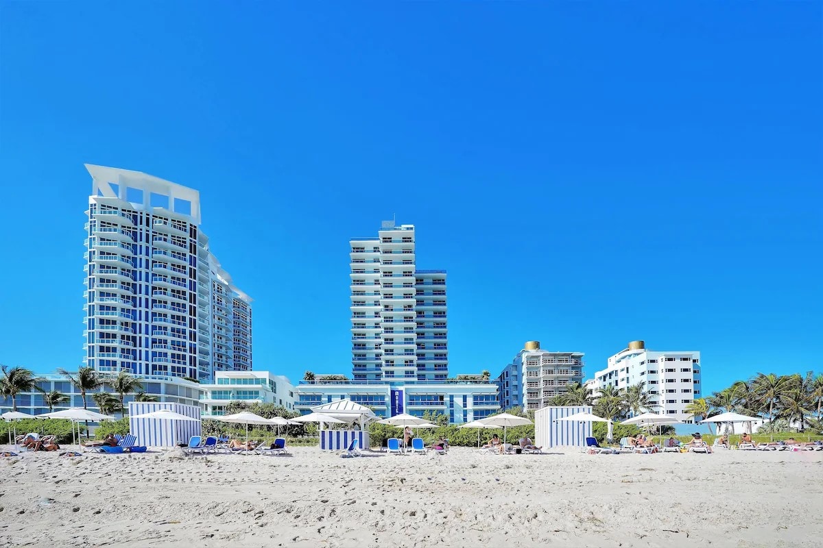 The image features a sandy beach with clear blue skies above. A row of beach umbrellas and loungers can be seen along the shoreline. Several tall buildings, including a high-rise and mid-rise structures, are situated in the background, reflecting a coastal vacation atmosphere.