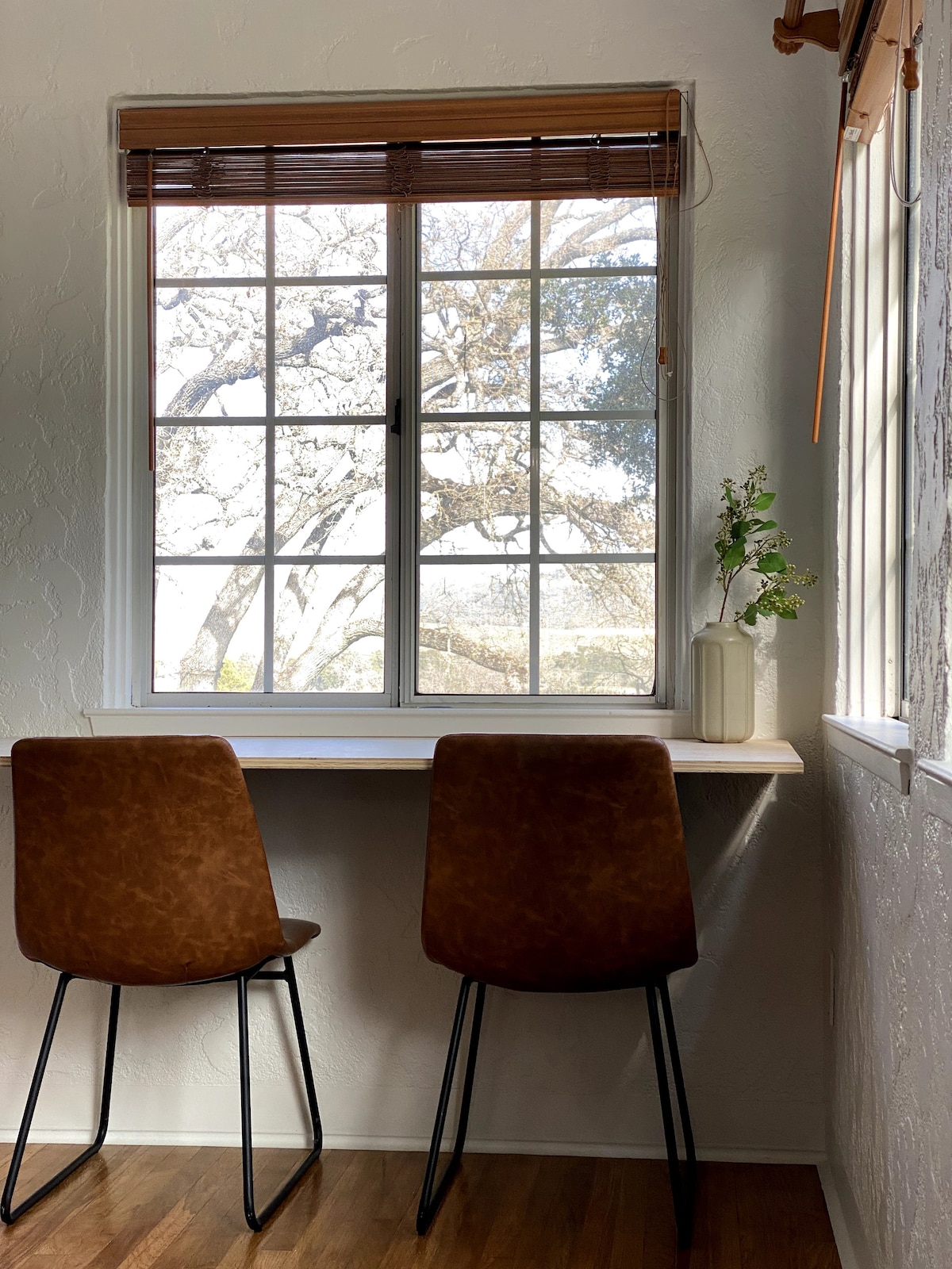 A cozy workspace is displayed with two brown chairs positioned at a light wood counter. A large window features multiple panes, framing views of oak trees and surrounding greenery. Natural light softly illuminates the space, enhancing the tranquil atmosphere.