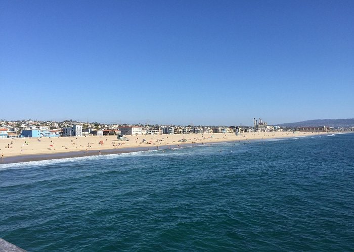 A wide view of the beach reveals soft sand meeting gentle waves, with numerous sunbathers scattered along the shore. Distant buildings line the coastline, offering a backdrop of urban life against the vibrant blue sky, complemented by clear water reflecting sunlight.