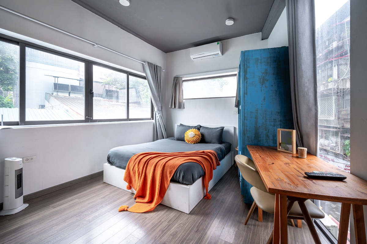 A spacious room features large windows allowing natural light to fill the space. A queen-size bed with grey bedding is complemented by a vibrant orange throw. A wooden table and chair provide a workspace, while an air conditioning unit is mounted on the wall.