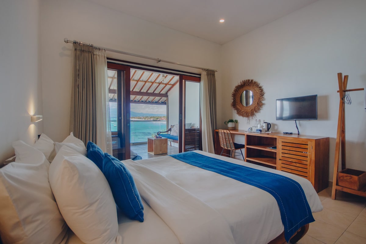 A spacious room features a comfortable bed adorned with white linens and a blue accent throw. Sliding glass doors open to a private balcony offering views of the sea. A wooden desk and chair sit against the wall, with a wall-mounted television nearby.