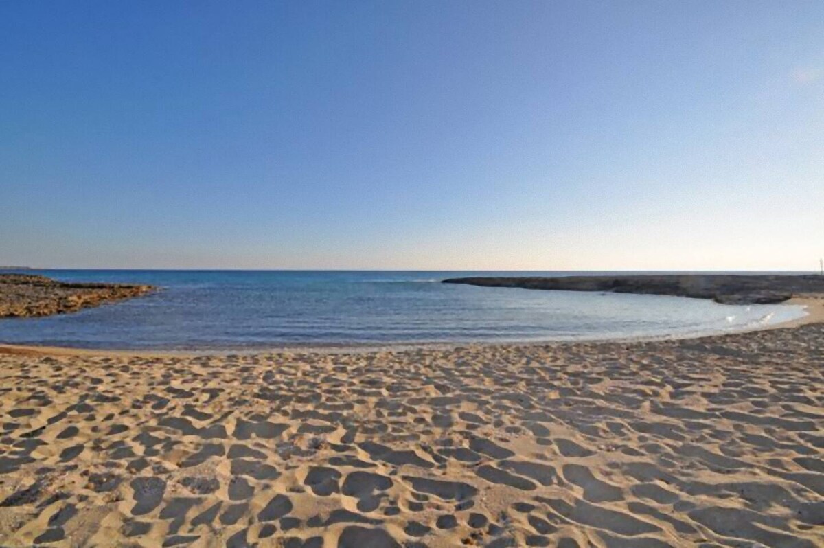 A serene beach scene features gentle waves lapping against a sandy shore. The clear blue water extends toward the horizon, under a bright sky. The soft, textured sand shows the outlines of footprints, while rocky formations can be seen along the water's edge.