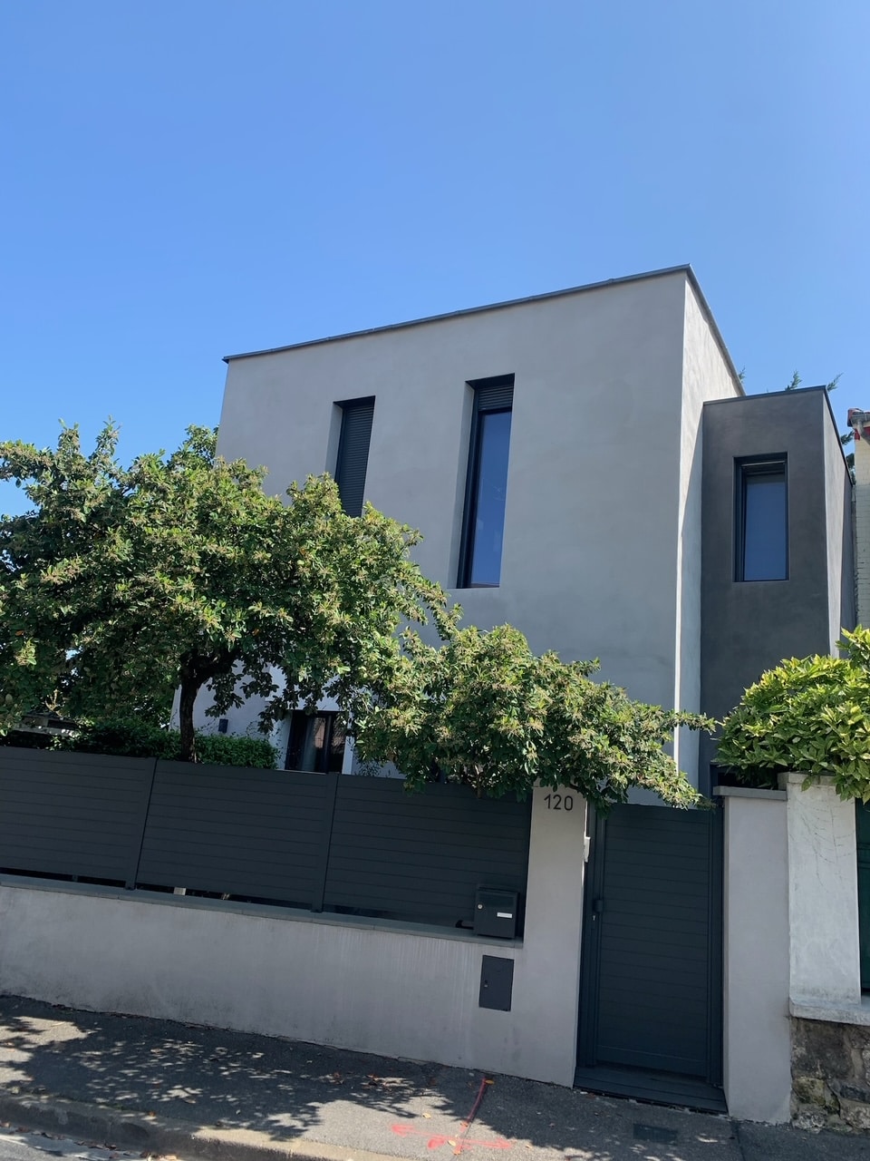 A modern, two-story house is showcased behind a sleek gray fence. The exterior features large, narrow windows and a minimalist design, complemented by surrounding green foliage. A well-maintained entrance door adds to the contemporary appeal, with clear blue skies overhead.