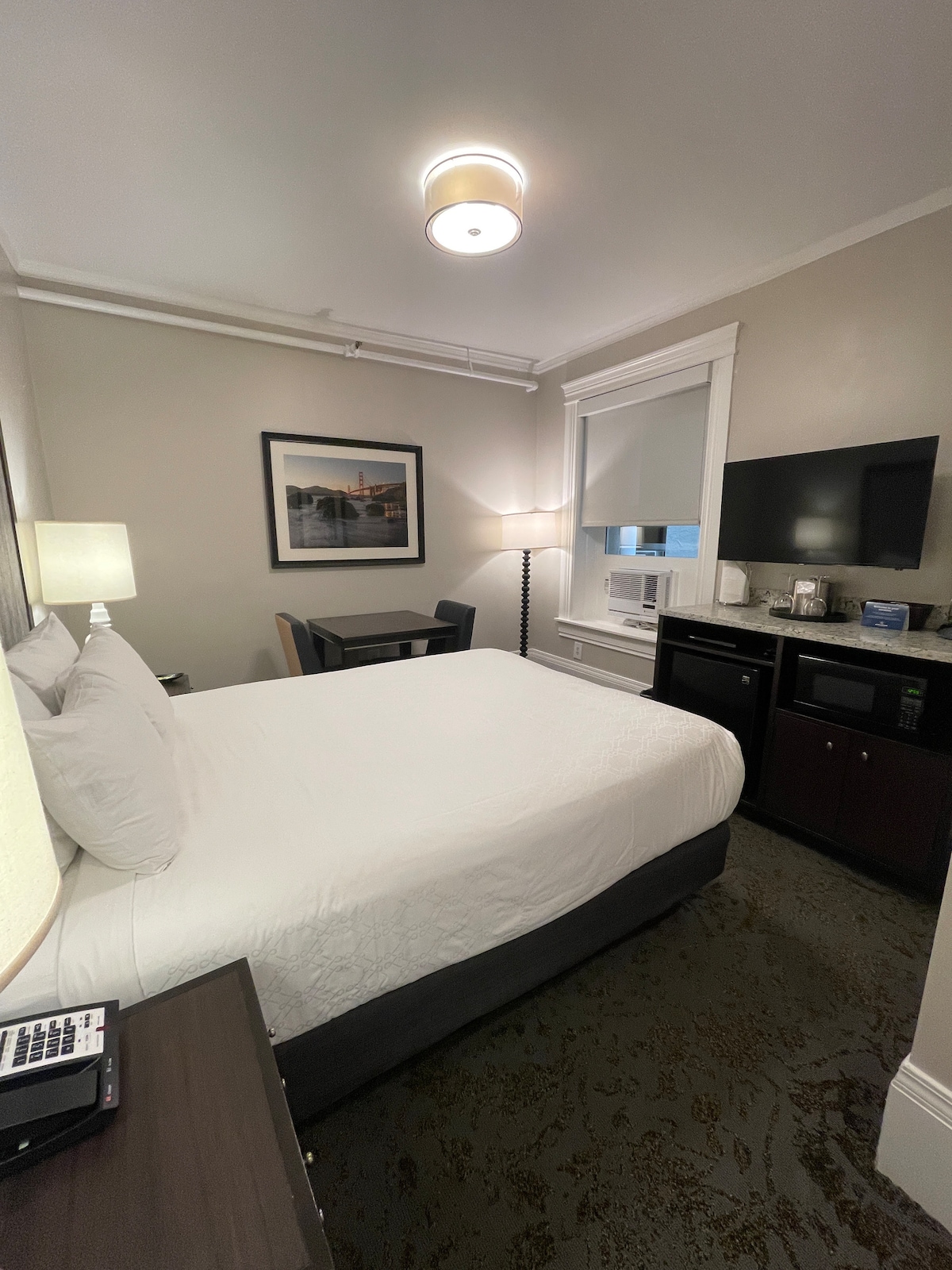 A well-appointed hotel room features a queen size bed with crisp white bedding. A small table and chair set is positioned near a window equipped with blinds. A flat-screen TV and a mini-fridge are situated on the opposite wall, contributing to the functional layout.
