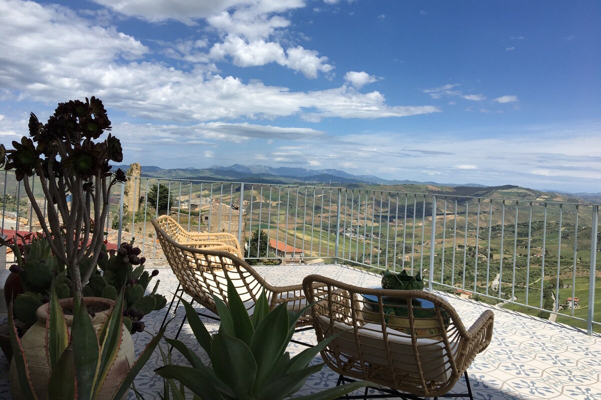 A terrace offers sweeping views of the surrounding hills and valleys, with a railing ensuring safety. Two comfortable chairs invite relaxation amidst greenery, while a table holds a potted succulent. The open sky is filled with scattered clouds, enhancing the scenic atmosphere.