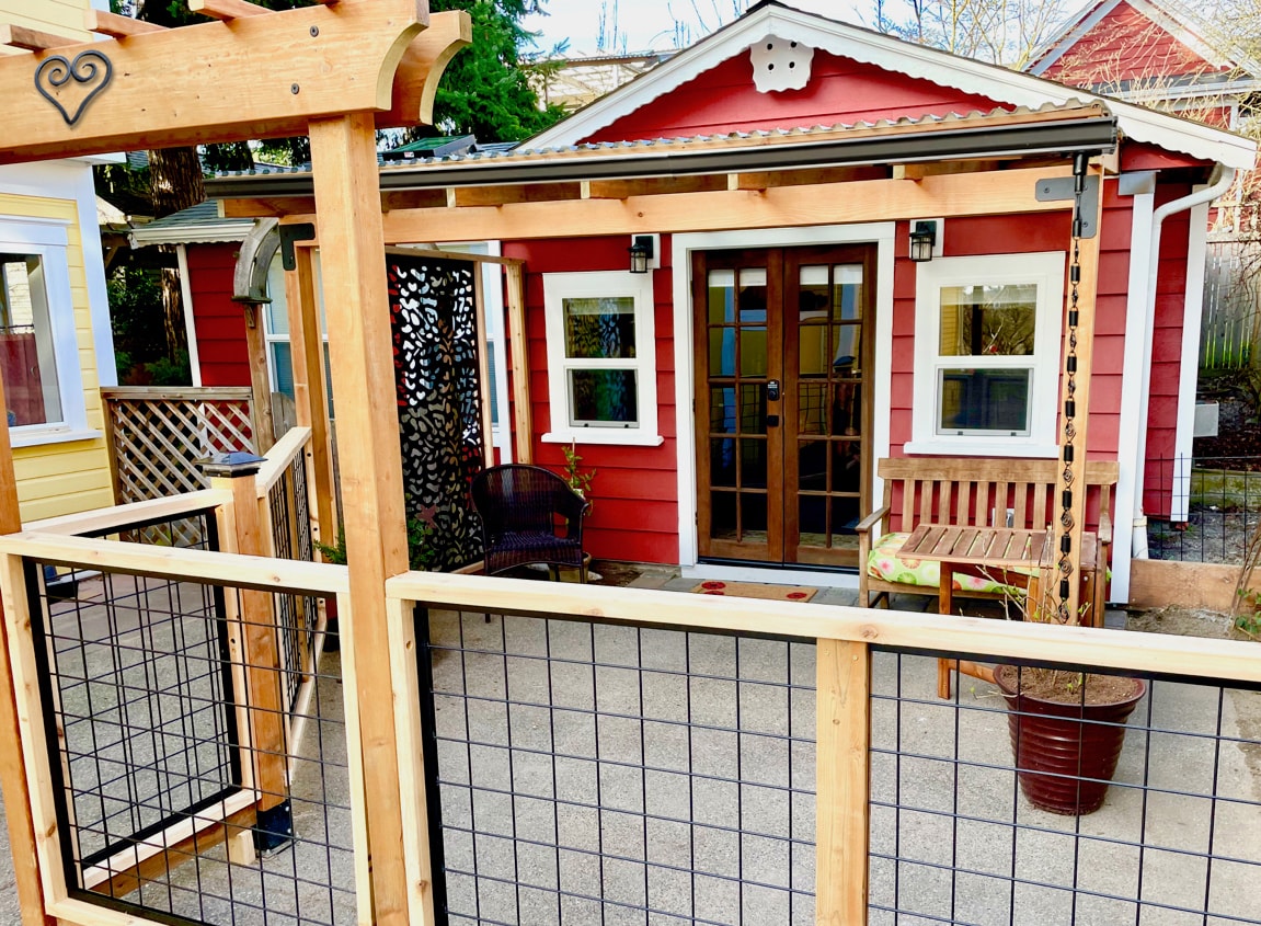 An inviting detached cottage is seen with a vibrant red exterior. Large glass doors framed by dark accents provide access to the interior. A fenced patio area includes a small bench and decorative plants, creating a welcoming outdoor space.