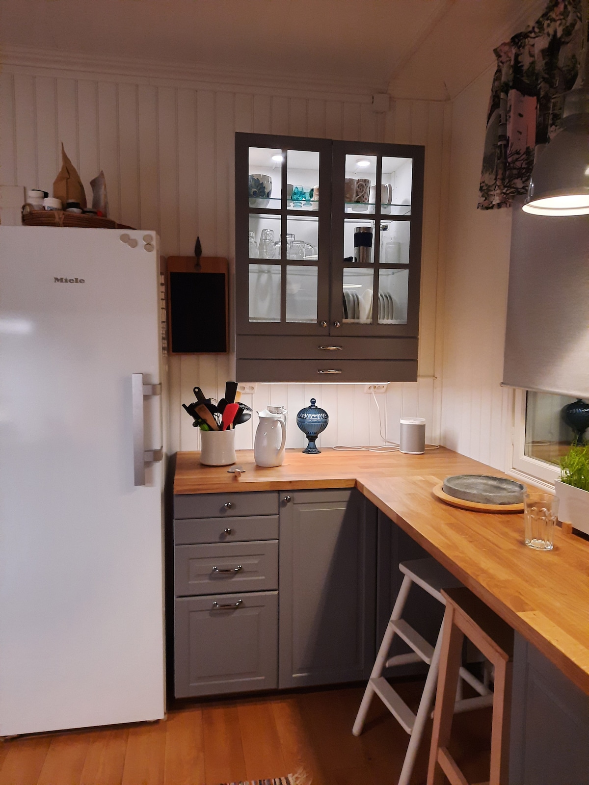 The kitchen features a white refrigerator and sleek gray cabinets. A wooden countertop is complemented by dishware displayed in a glass-front cabinet. Cooking utensils are organized neatly. Natural light illuminates the workspace, highlighting the clean lines and functional design.