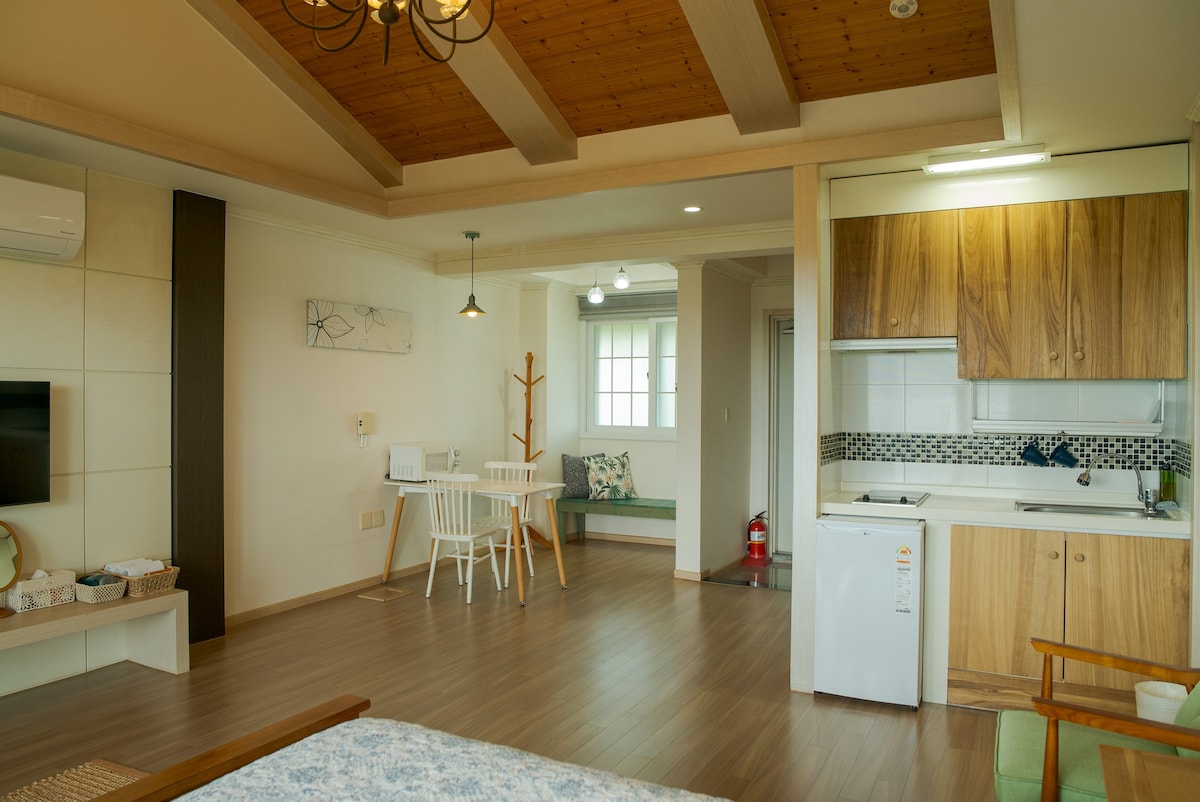 An open living space features wooden beams on the ceiling and a cozy kitchen area. A dining table with four chairs is visible, along with a small sofa in the corner. Natural light brightens the room through a window, complementing the warm tones of the furnishings.