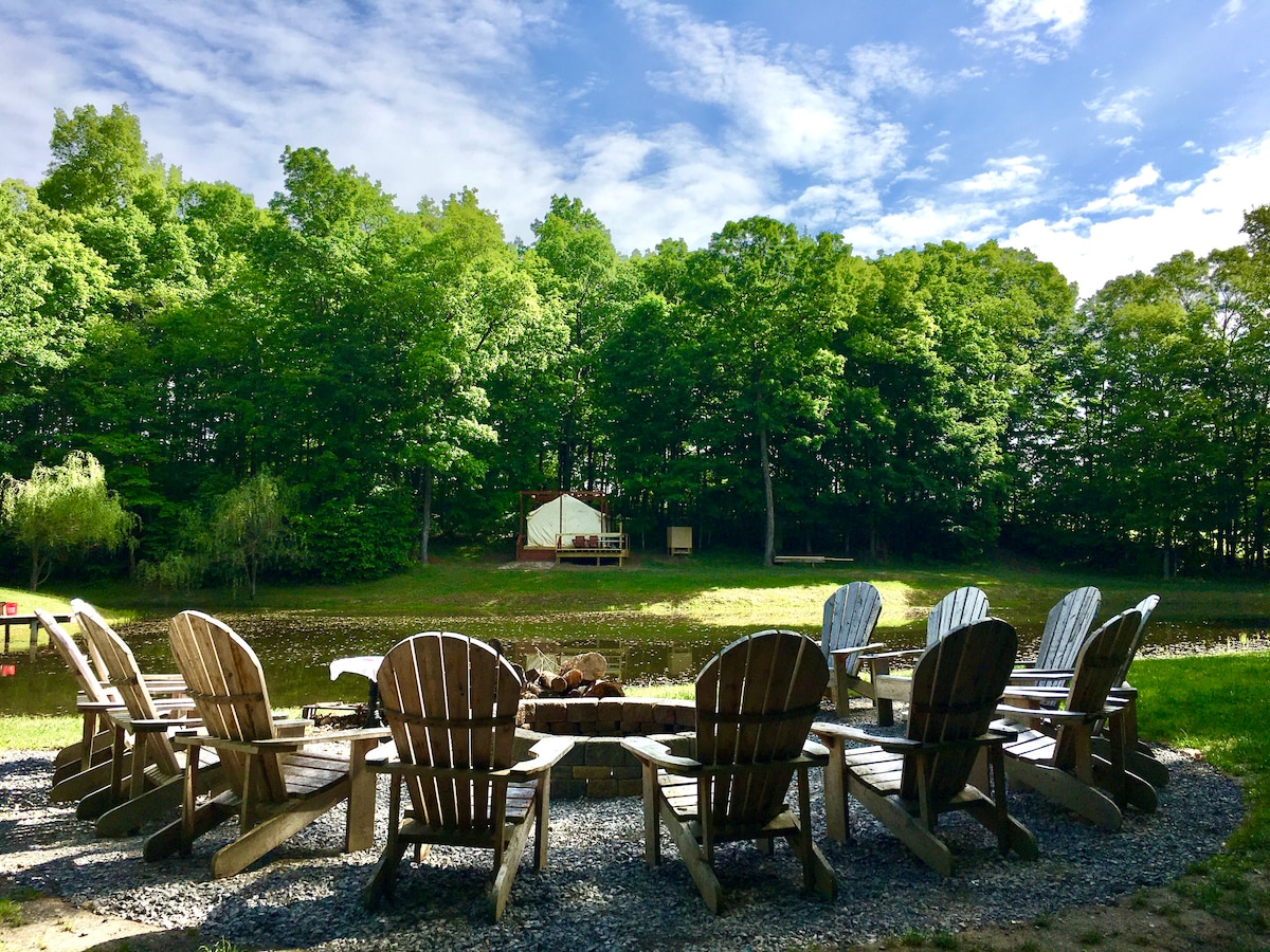 A circle of wooden Adirondack chairs surrounds a stone fire pit, situated on a gravel area. In the background, a boutique tent is visible near a tranquil koi pond, framed by lush green trees under a partly cloudy sky.