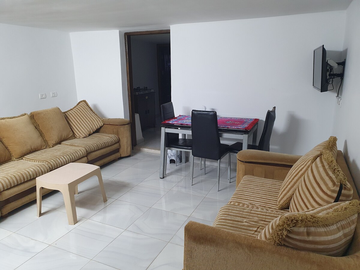 The living area features a spacious arrangement of light brown sofas with striped patterns, accompanied by a simple coffee table. A dining table with black chairs is positioned nearby, complemented by a television mounted on the wall and illuminated by natural light.