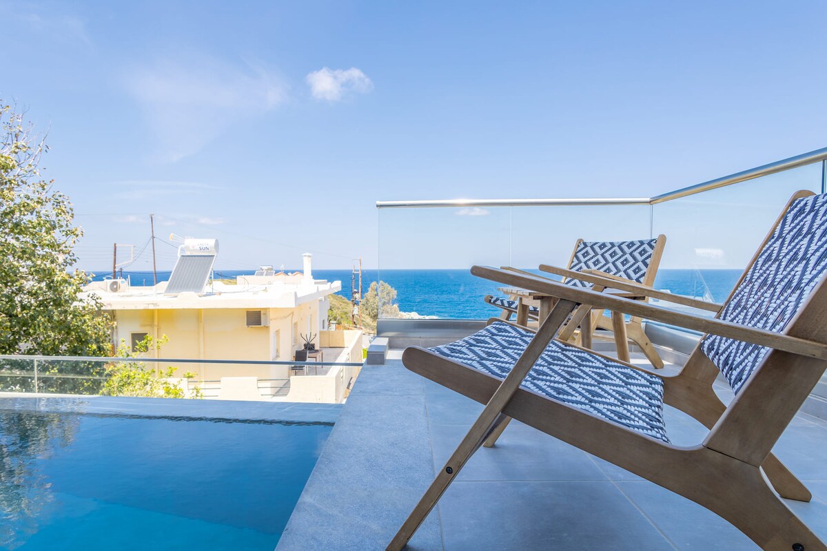 A serene outdoor area features comfortable wooden lounge chairs with patterned cushions. The private swimming pool reflects the clear blue sky, while the expansive view of the ocean is visible in the background, enhancing the space's relaxing ambiance.