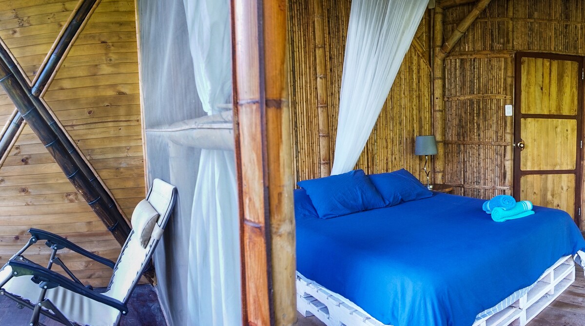 The cozy bedroom features a spacious bed dressed in soft blue linens and surrounded by bamboo walls. A relaxing chair sits near the entrance, and natural light illuminates the space through a window adorned with sheer curtains.