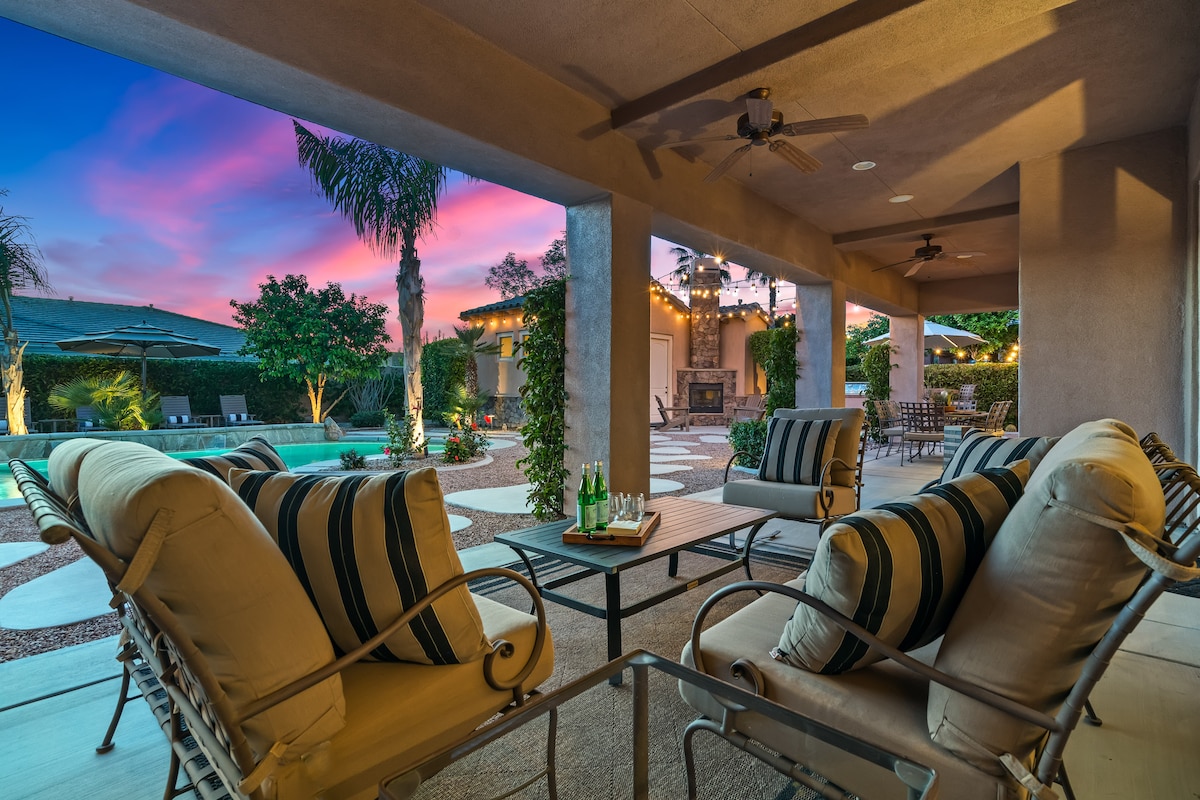 An outdoor living area features comfortable seating with striped cushions and a central table. The space is framed by columns and offers views of a softly lit garden and an inviting pool. A vibrant sunset sky fills the background, enhancing the tranquil ambiance.