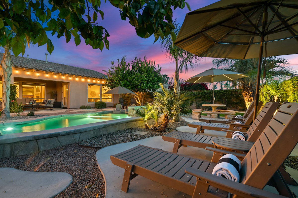 The outdoor area features a serene pool surrounded by lounge chairs and umbrellas. Lush landscaping and palm trees provide privacy, while string lights emit a warm glow, enhancing the inviting atmosphere. The sky showcases vibrant colors at dusk, adding to the tranquil setting.