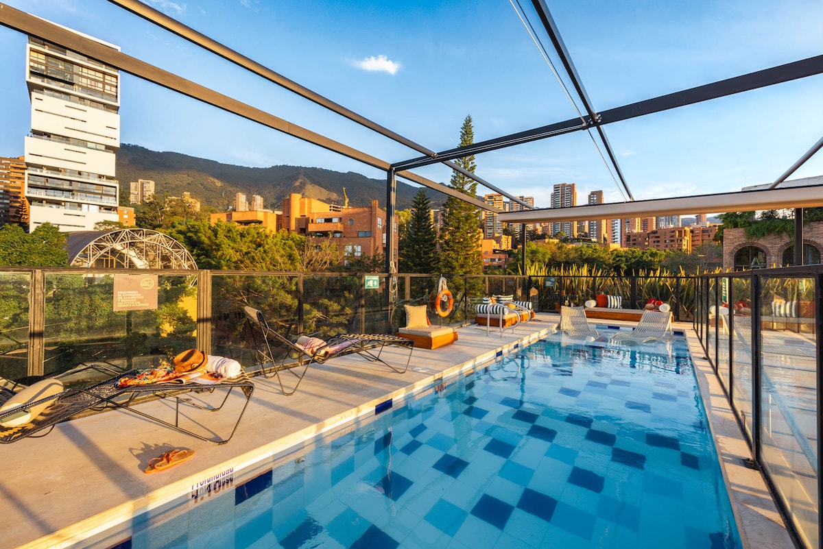 An outdoor pool area is showcased, framed by glass railings and a modern canopy. Lounge chairs are positioned around the pool, offering spaces for relaxation. Lush greenery and city views are visible in the background, with mountains rising in the distance under a clear blue sky.