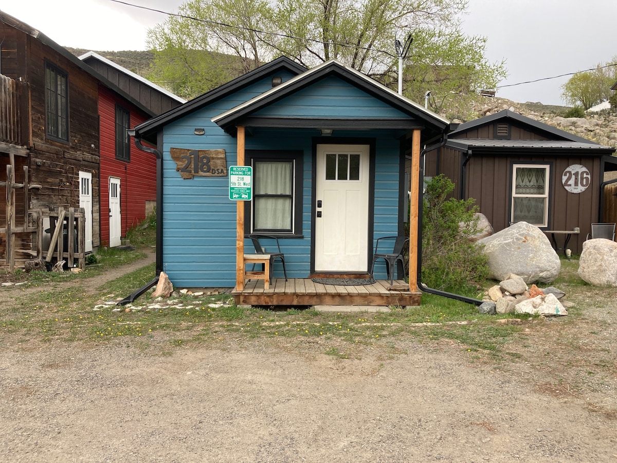The cabin's exterior features a blue façade and a welcoming front porch, complete with two chairs. Surrounding natural elements include grassy areas and stones, while neighboring buildings are visible in muted colors, contributing to an urban yet quaint atmosphere.