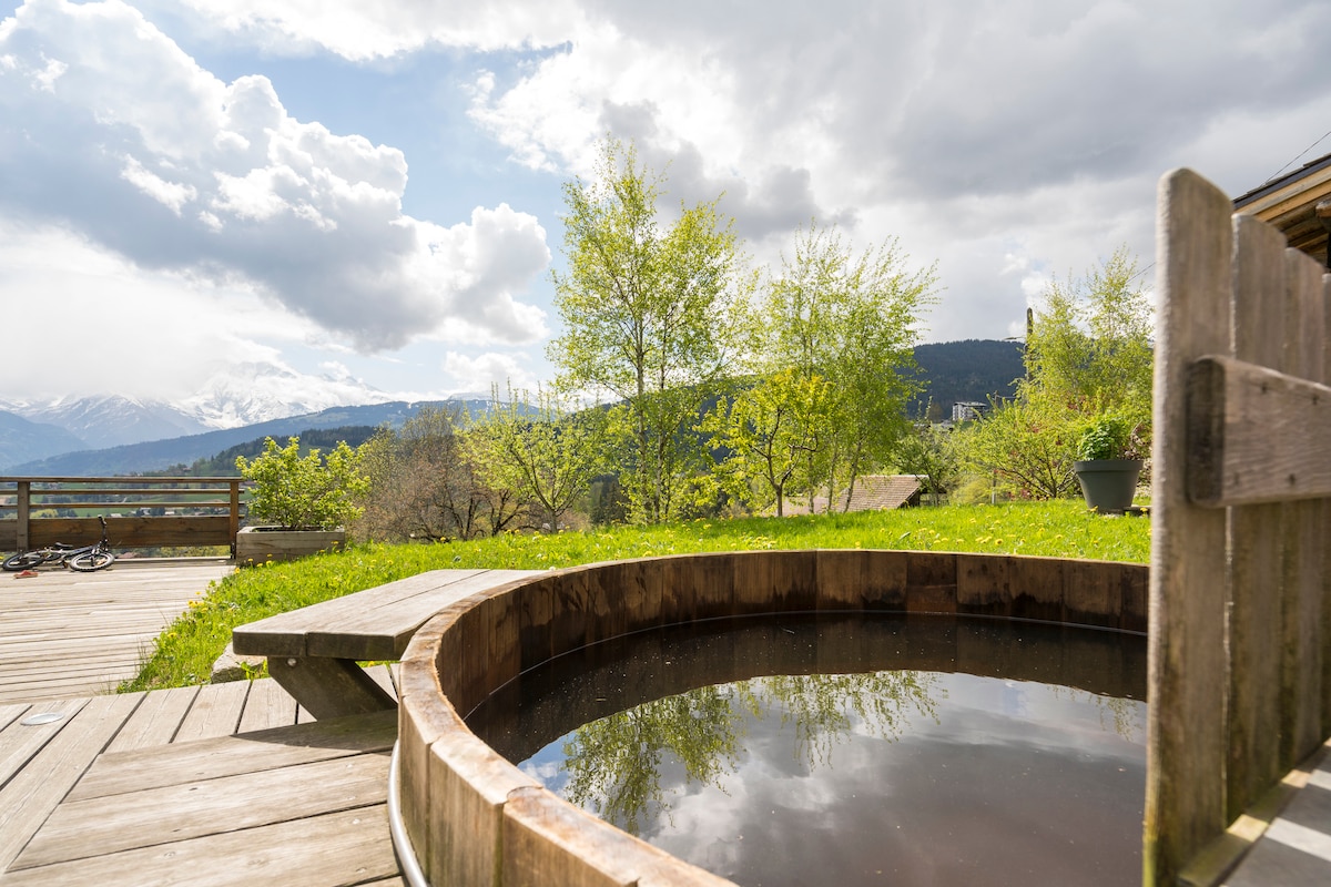 A wooden spa surrounds a tranquil pool, situated on a spacious deck. Lush greenery and trees are visible in the background, providing a serene atmosphere. The distant mountains, including Mont Blanc, enhance the view, under a partly cloudy sky.