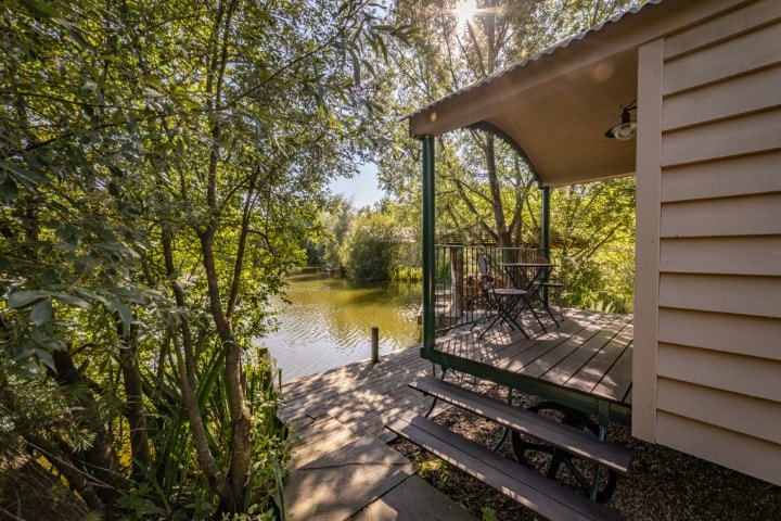 A serene outdoor deck area is visible, featuring a small table and two chairs overlooking a peaceful lake. Lush greenery surrounds the space, creating a natural and tranquil setting.