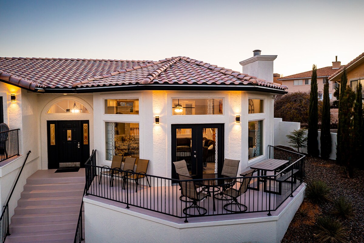 An exterior view of the vacation home is presented, showcasing a spacious deck with seating for six. The building features large windows allowing natural light to illuminate the interior. Warm lighting from the porch lamps adds a welcoming touch to the structure's design.