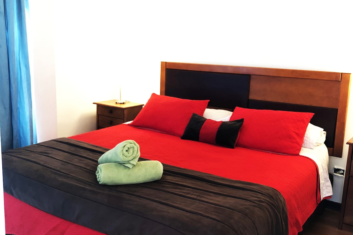 A spacious bed is showcased with a red and brown coverlet, complemented by two red pillows and a black accent pillow. Neatly rolled green towels are placed at the foot of the bed. Light enters through a curtain-covered window, creating a bright ambiance.