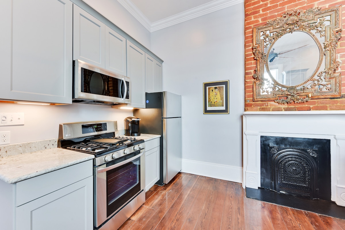 A modern kitchen features stainless steel appliances, including a gas stove and refrigerator. Light grey cabinetry contrasts with the warm brick wall and intricately framed mirror. A granite countertop provides ample workspace, while the elegant fireplace adds a classic touch to the room.