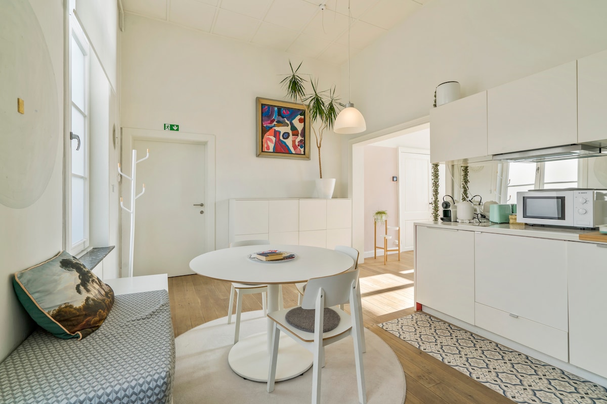 A light-filled dining area features a round table surrounded by three chairs, complemented by a cozy seating nook. A modern kitchen is visible in the background, equipped with a microwave, and adorned with tasteful artwork and plants that enhance the contemporary design.