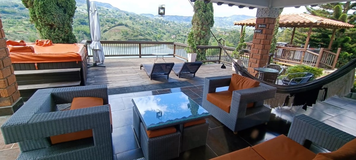 An outdoor terrace features a lounge area with two sun chairs and a coffee table, surrounded by green hills. A hammock is visible in the background, providing space for relaxation. The wooden deck extends to a stunning view of the landscape and water.