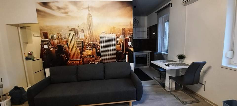 A modern living area features a large mural of a city skyline, creating a vibrant backdrop. A dark sofa is positioned in front, while a dining table with gray chairs is visible in the background. Natural light streams through a nearby window.