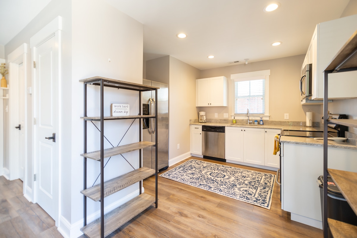 A modern kitchen is presented with white cabinets and stainless-steel appliances, including a refrigerator and dishwasher. A large window allows natural light to brighten the space. The area features a decorative rug on the wooden floor and open shelving for additional storage.