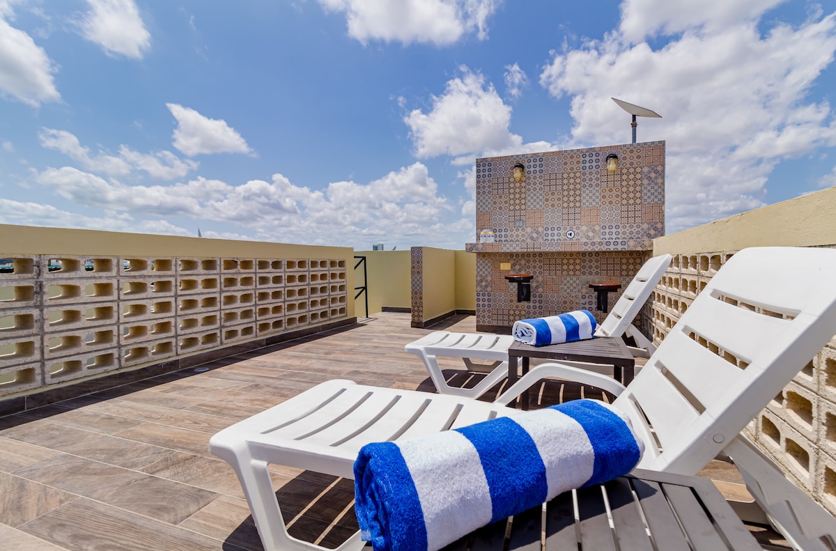 A spacious rooftop terrace is shown with two lounge chairs, each adorned with a striped towel. The area features a tiled wall and a small table, all under a partly cloudy sky, providing a relaxing space for guests.