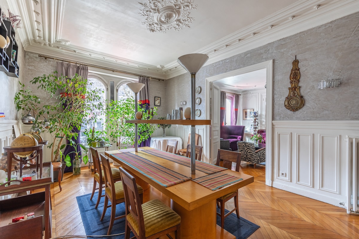 A spacious dining area features a large wooden table surrounded by eight chairs, complemented by a patterned runner. The room is enhanced by large windows allowing natural light in, with decorative plants that add a touch of greenery to the space.