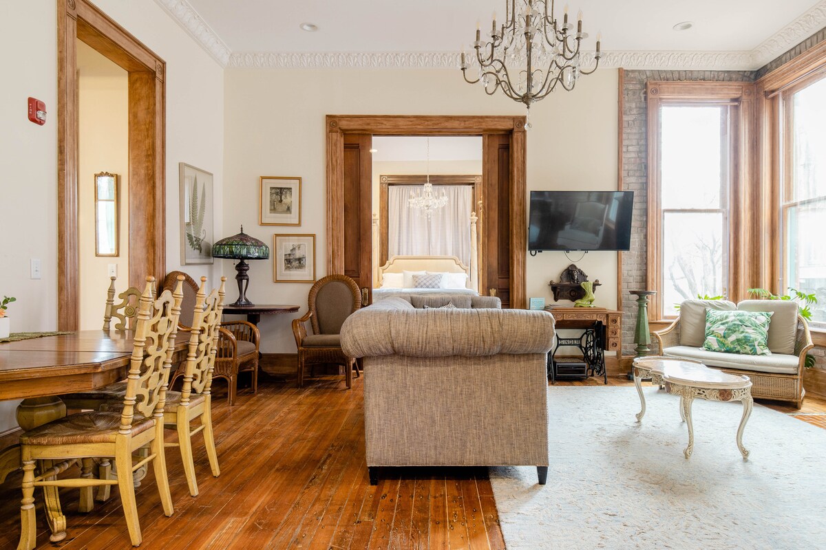 A spacious living area features tall windows allowing natural light to fill the room. Elegant furnishings, including a plush sofa and ornate coffee table, are arranged around a warm, wooden floor. A chandelier hangs from the ceiling, adding a touch of sophistication, while a dining table is positioned nearby.