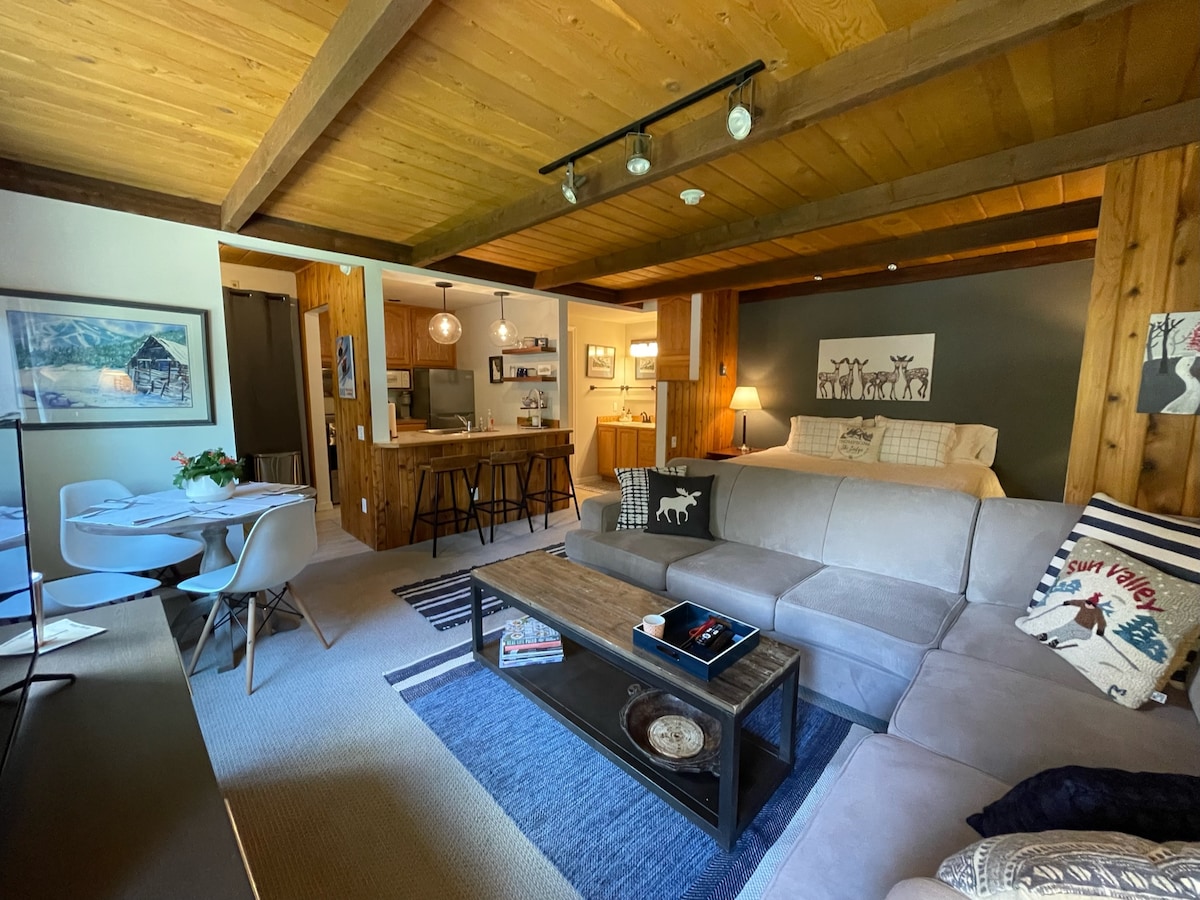 A cozy living area features a spacious sectional sofa and a coffee table, complemented by warm wood beams and a stylish rug. In the background, an open kitchen with bar seating and a dining table creates an inviting atmosphere. Natural light highlights the decor.