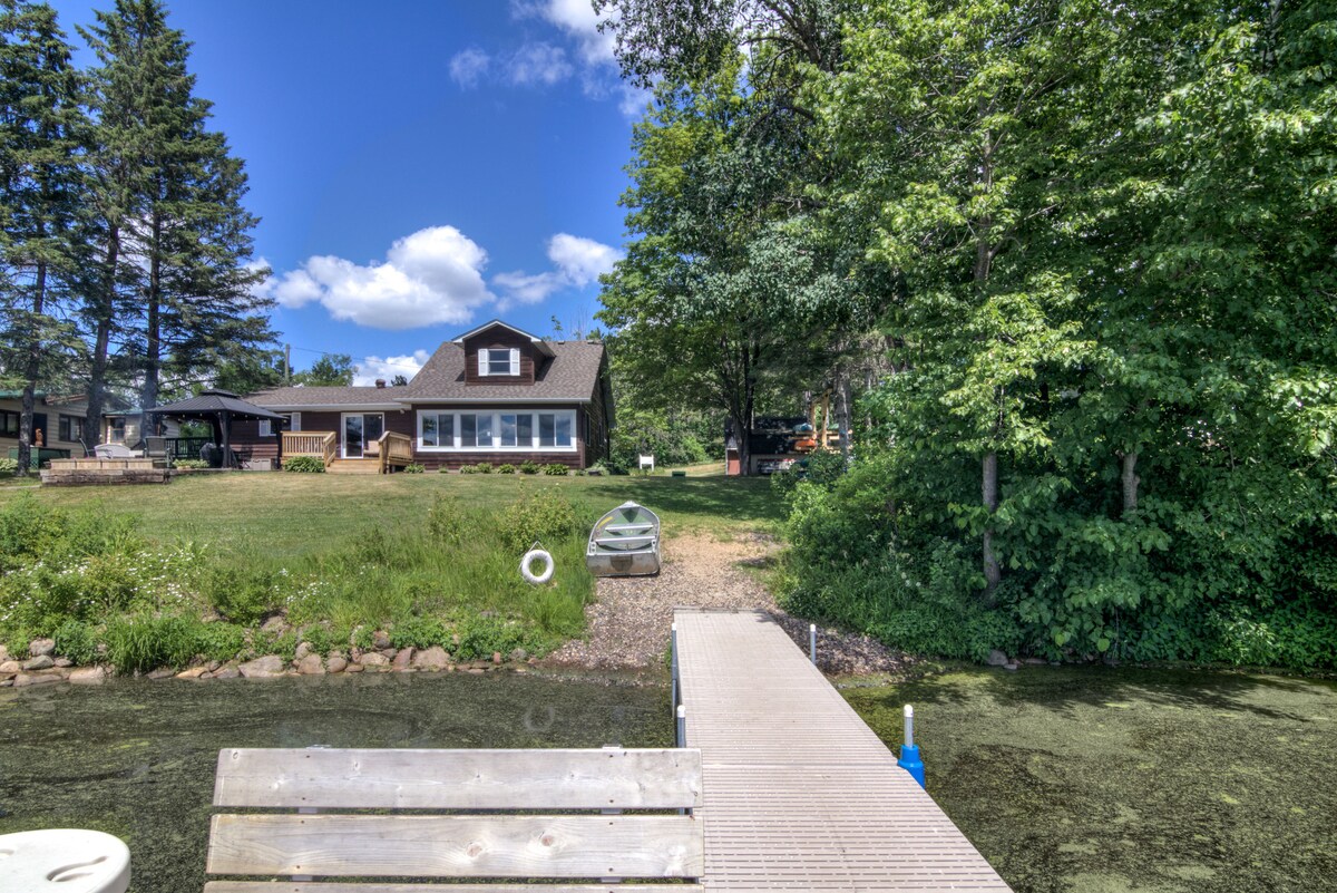 A spacious yard leads to the private dock, where a canoe is visible on the grassy shore. The cabin, framed by tall trees, features large windows that reflect the sky. A cozy sitting area is noticed near the fire pit, inviting relaxation by the river.