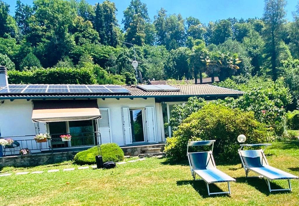 A private villa is set amidst a lush green landscape, featuring an expansive garden with sun loungers arranged invitingly under clear skies. Solar panels are installed on the roof, reflecting a focus on sustainability. The home's large windows create an airy connection with the outdoors.