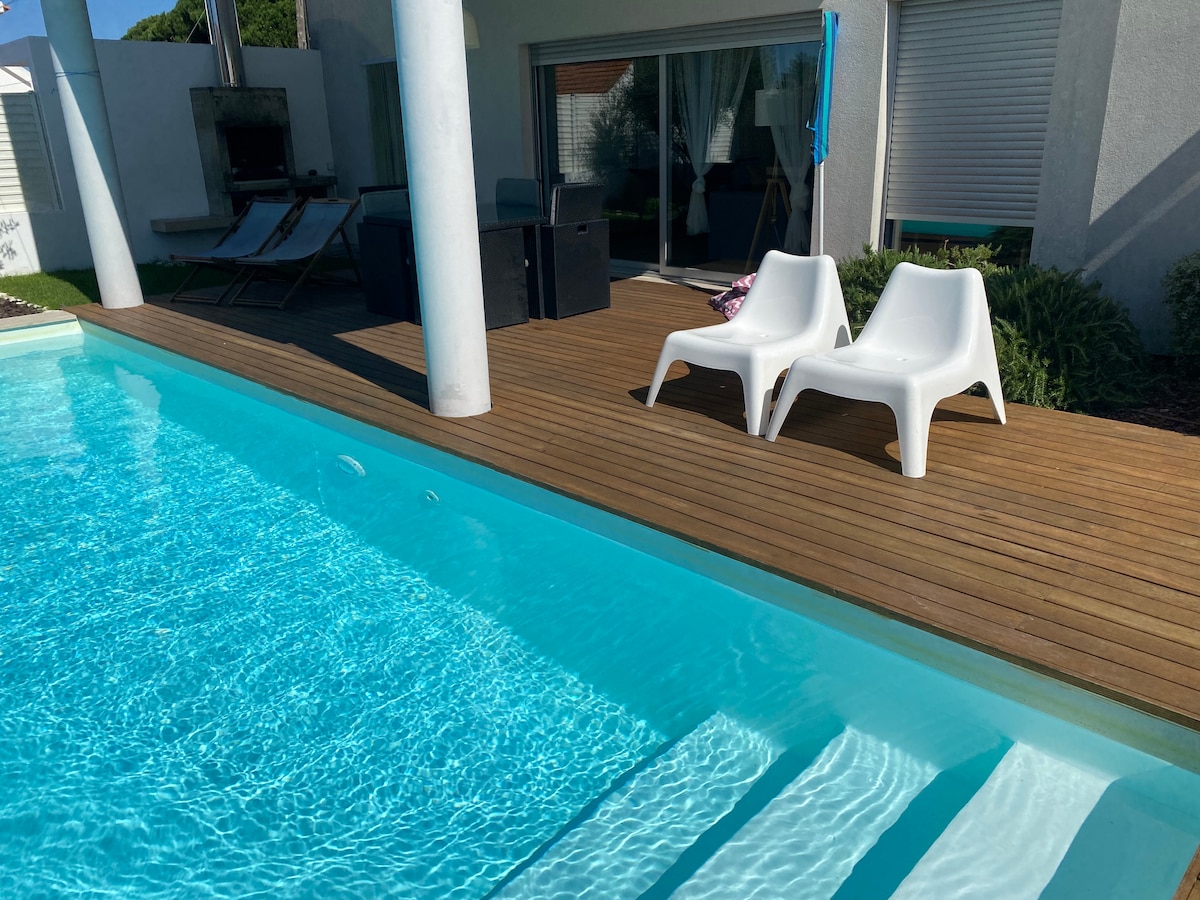 A private pool glimmers under sunlight, with two white lounge chairs positioned beside it on a wooden deck. The pool steps invite entry, while sliding glass doors provide visibility into a modern living space in the background.