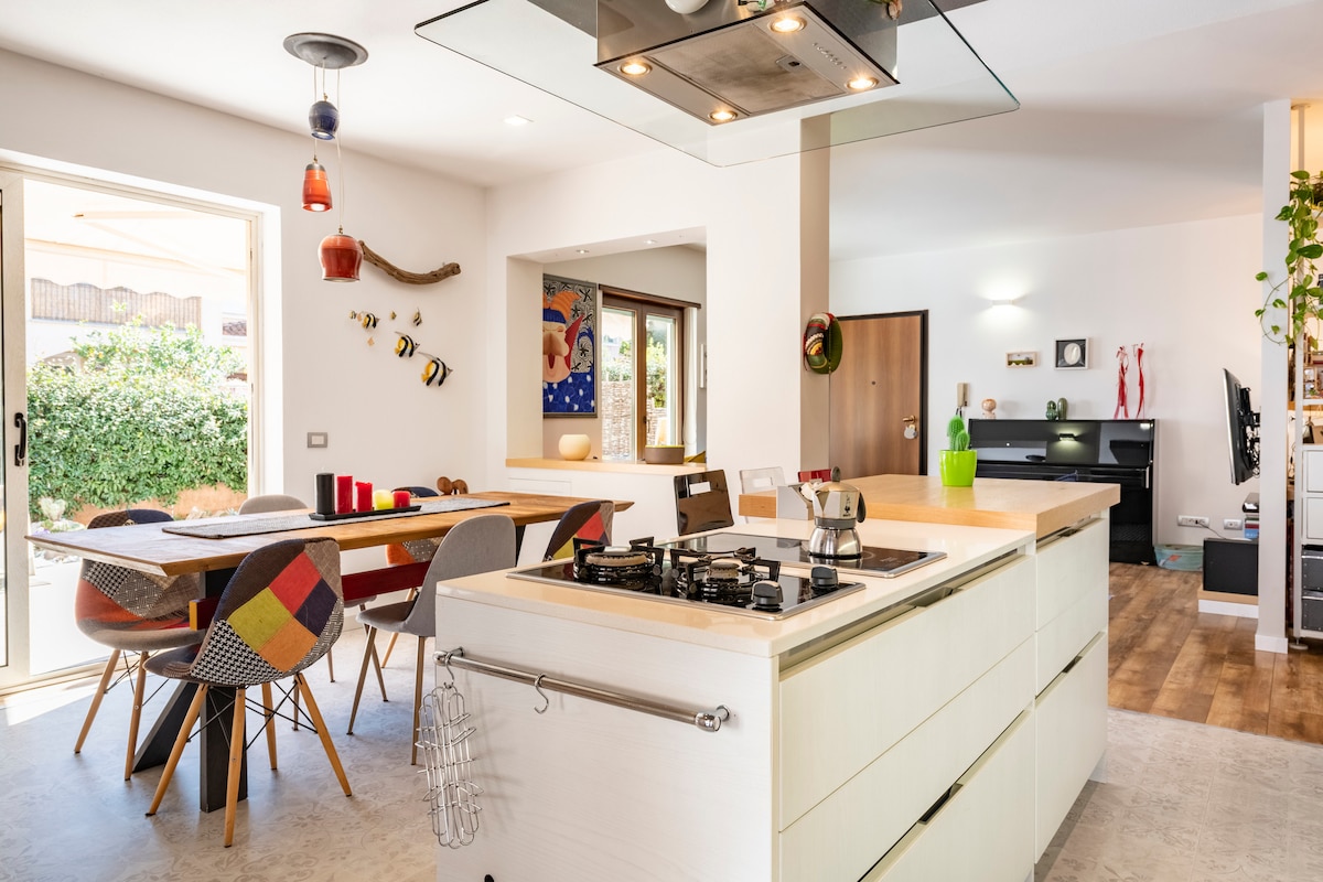 A modern kitchen area features an island with a gas stovetop, providing a central cooking space. Colorful dining chairs surround a wooden table, while large windows allow natural light to illuminate the room. A view into the garden is visible through the glass door.