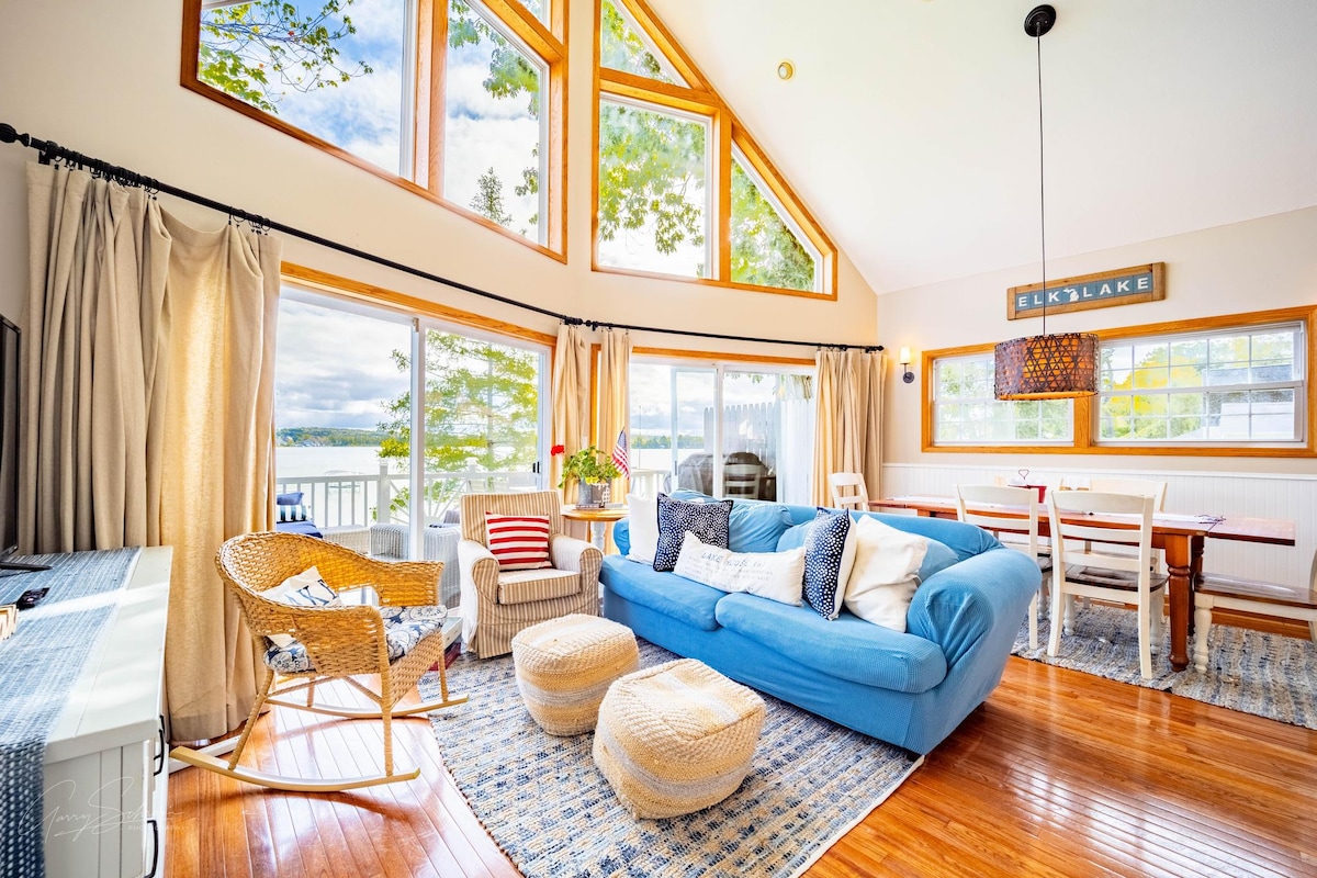 A sunlit living area features large windows offering views of Elk Lake. A blue sofa is complemented by two woven ottomans and a rattan chair. A dining table with white chairs sits nearby, with light white curtains framing the windows, creating a bright and airy atmosphere.