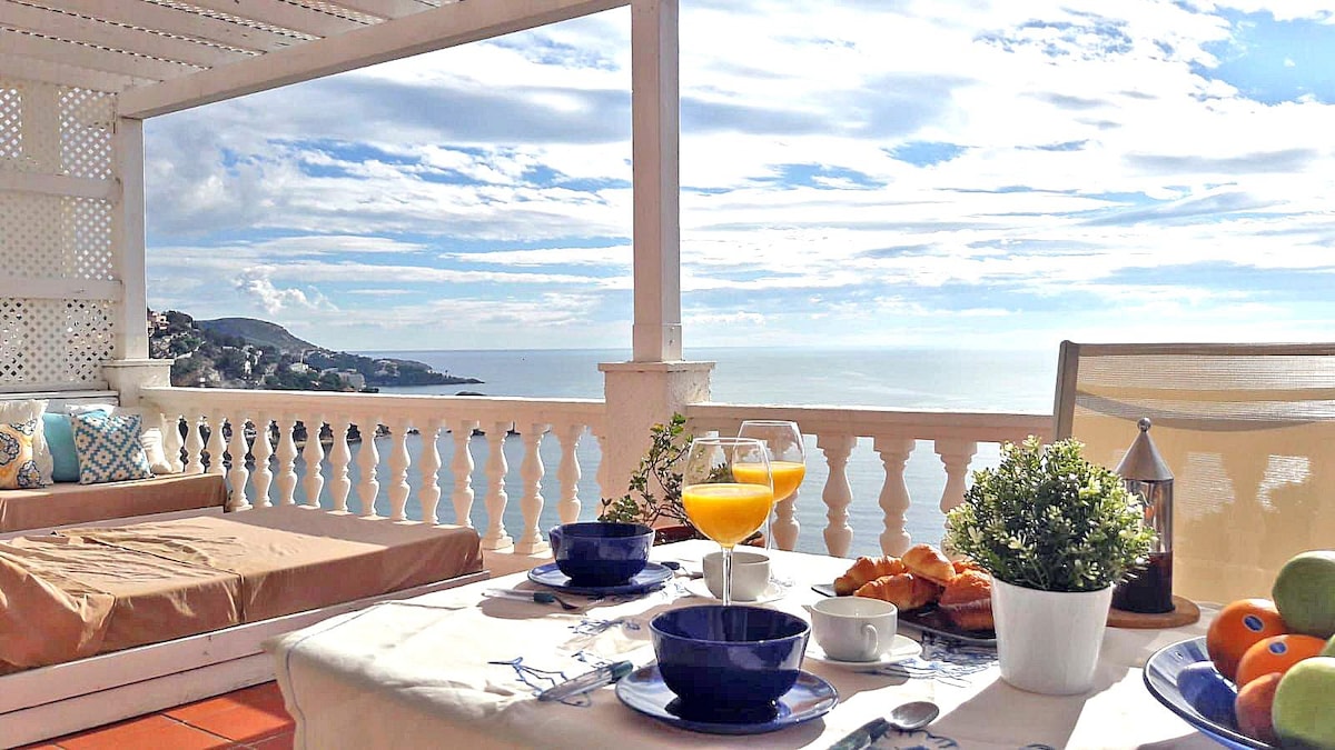 An outdoor terrace features a dining table set for breakfast with a view of the sea. A glass of orange juice, a bowl of fruit, and pastries are arranged on the table. A cozy lounge area with a sofa offers a relaxing spot to enjoy the scenery.