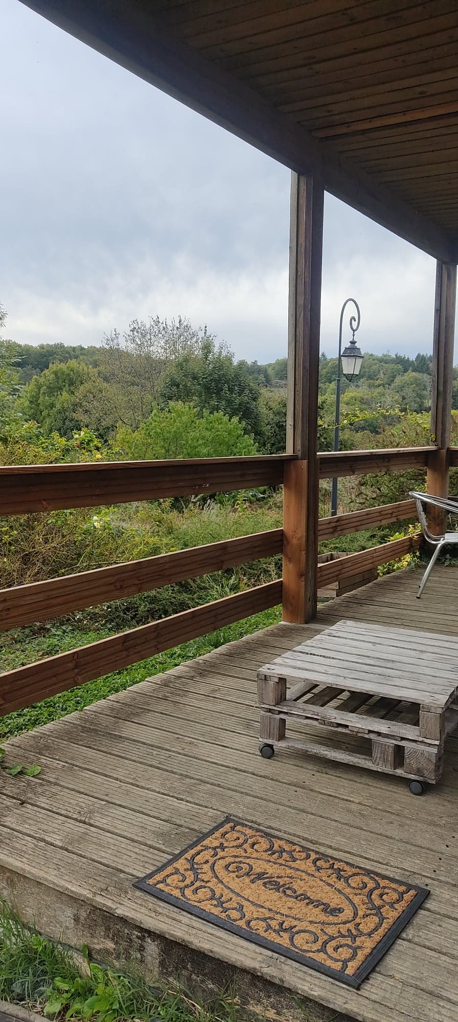 An outdoor deck is framed by wooden railings, providing views of lush greenery and distant hills. A small table made from pallets rests on the wooden floor alongside a simple metal chair. A vintage lamp post adds a touch of character to the serene landscape.