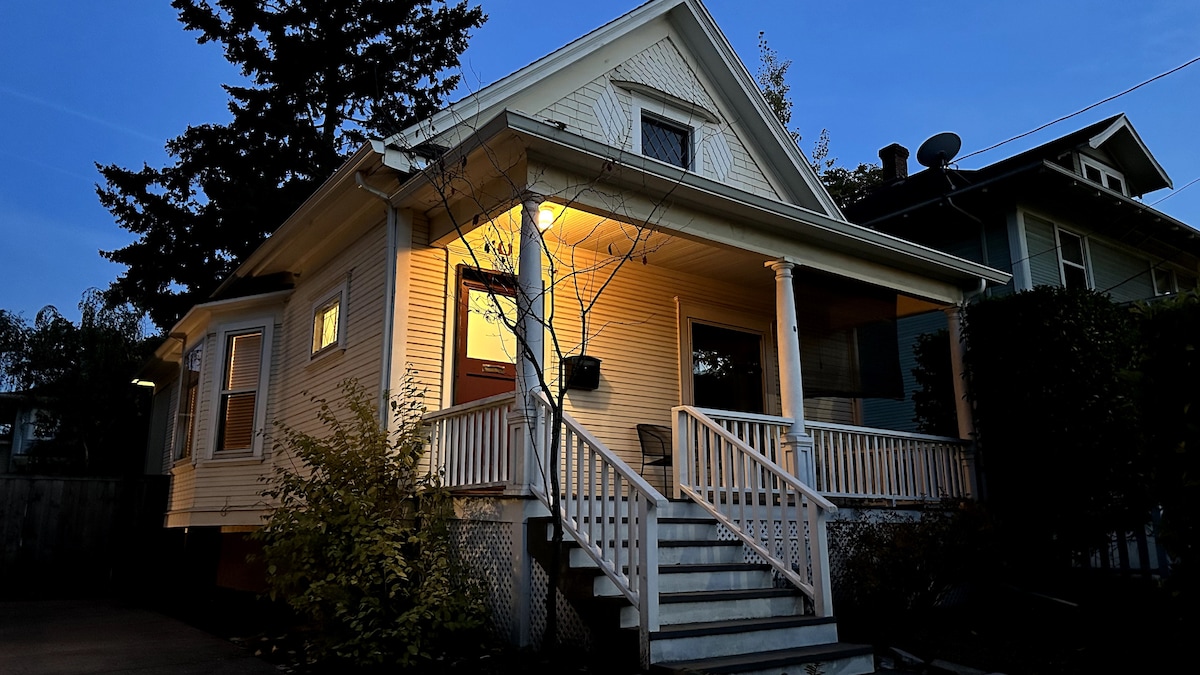 The exterior of a cozy vintage home is shown, featuring a welcoming porch with white railings and steps leading to the entrance. Soft lighting highlights the faΓ§ade, while surrounding trees add a touch of nature. The neighborhood street can be seen quietly in the background.