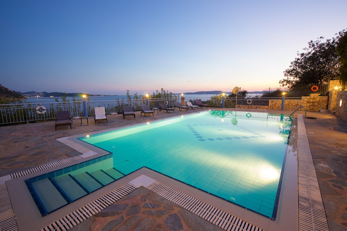 A serene outdoor pool area is presented, featuring clear turquoise water reflecting the evening sky. Lounge chairs are positioned around the pool, with views of the sea visible in the background. Soft lighting adds to the tranquil atmosphere as daylight fades.
