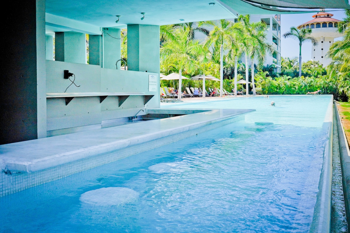 A serene pool area is viewed, featuring a long, shallow swimming pool bordered by tropical palm trees. A bar counter extends along the pool edge, with submerged seating stones incorporated into the water, providing a space for relaxation and enjoyment.