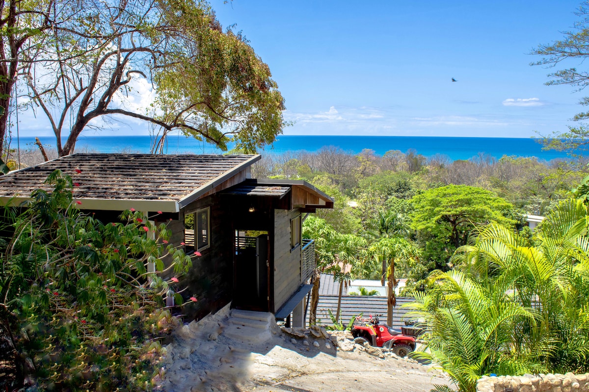 The bungalow sits elevated on a hillside, with a view of the vibrant blue ocean in the distance. Lush greenery surrounds the entrance pathway, which leads to a welcoming door. A vehicle can be seen parked nearby under bright sunlight.